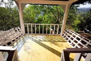 A covered outdoor area with a wooden deck, white railings, and a view of a forested landscape. - Temple Garden (Canacona)