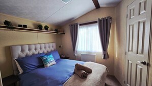 Un dormitorio con cabecera capitonada, cama con ropa de cama azul, almohada con el texto 'Ocean Vibes', una toalla enrollada, estante con plantas y ventana con cortinas.