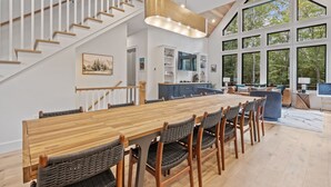 Un'ampia sala da pranzo con un lungo tavolo in legno e sedie in vimini, una scala con ringhiere in legno e ampie finestre che si affacciano su alberi.
