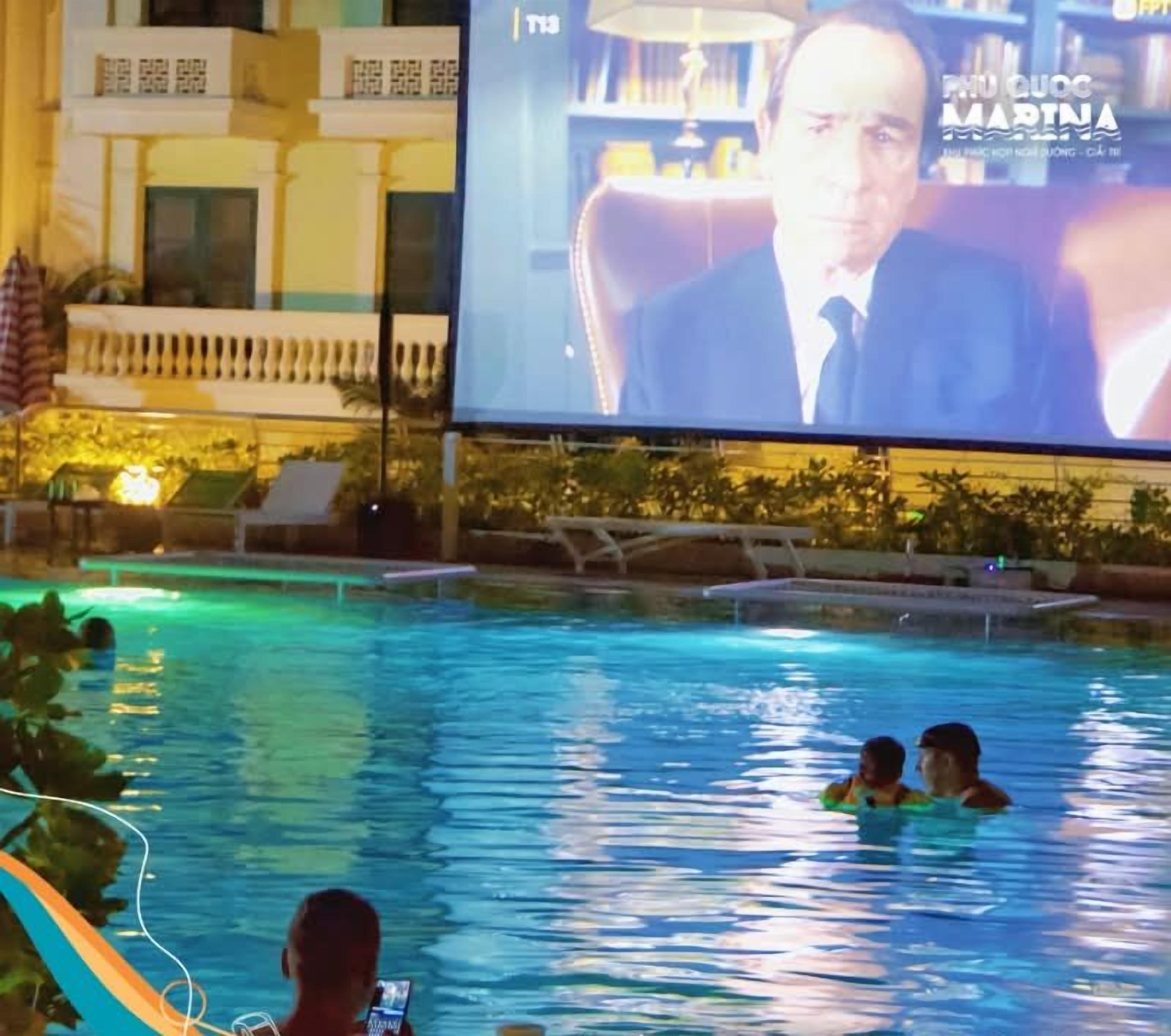 A swimming pool area with lounge chairs and a large screen displaying a news broadcast.