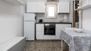 Een kleine keuken met witte kasten, een ingebouwde oven, een raam boven de gootsteen en een tafel met een tafelkleed met dessin.