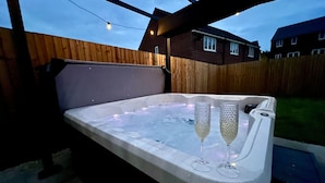 A hot tub with two glasses of champagne, surrounded by a wooden fence and houses in the background.