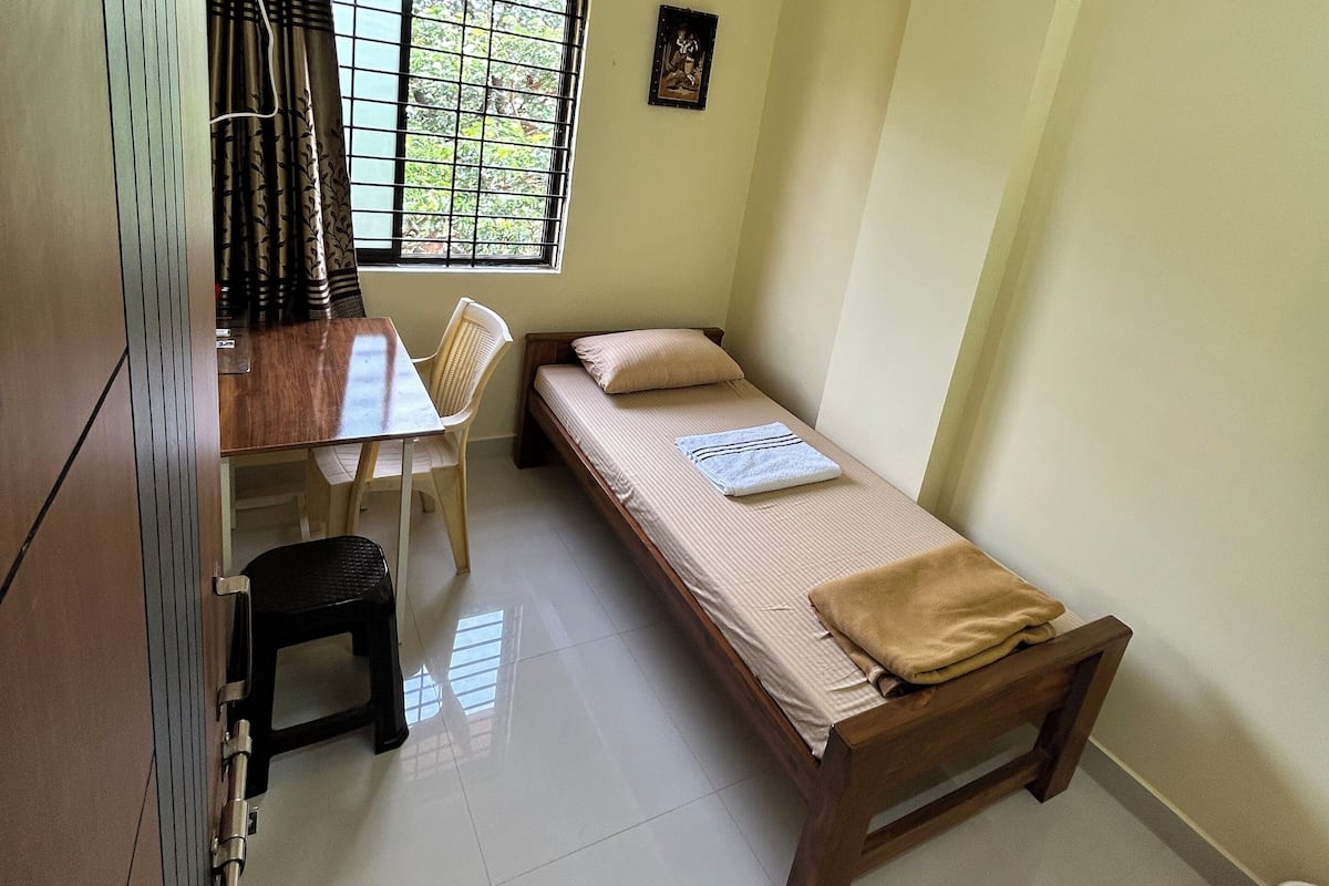 A small, tidy room with a bed, a desk, and a chair. There's a window with bars, a wall-mounted fan, and a picture frame.
