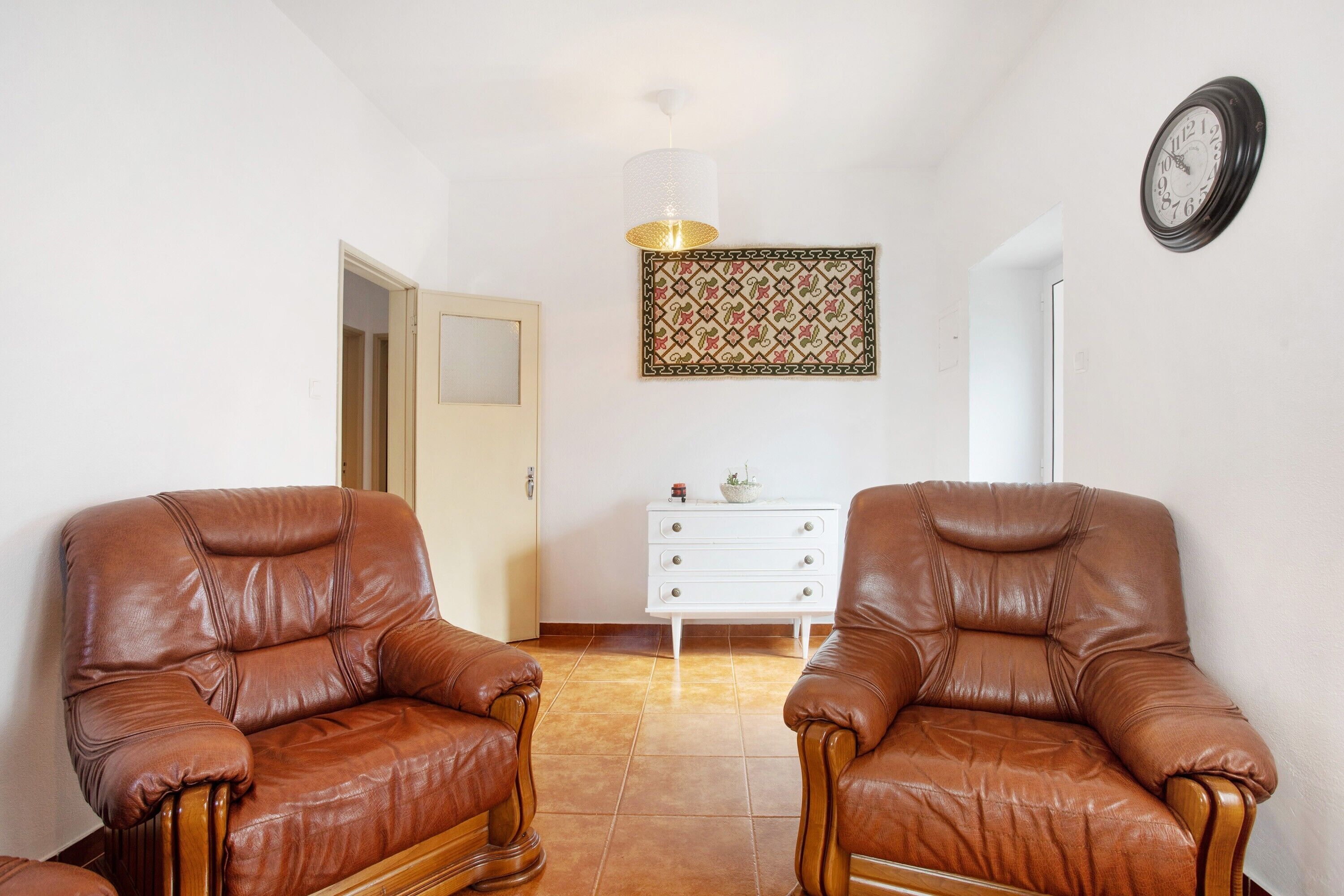 Una habitación con dos sillones de cuero, un tocador blanco, un tapiz decorativo y un reloj de pared.