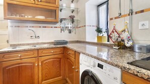 A kitchen with wooden cabinets, a granite countertop, a built-in washing machine, and a glass shelf with kitchen utensils. - Holiday Home 'D´Aldeia' with Shared Terrace, Private Garden and Wi-Fi (Cela Velha)