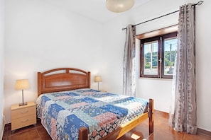 Quarto com cama de madeira, mesa de cabeceira com abajur, janela com cortinas e vista para o verde lá fora.