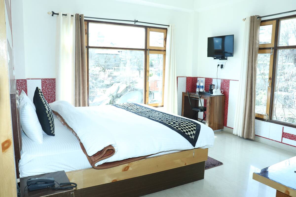 A hotel room with a large bed, a desk with a chair, a television mounted on the wall, and large windows with curtains.