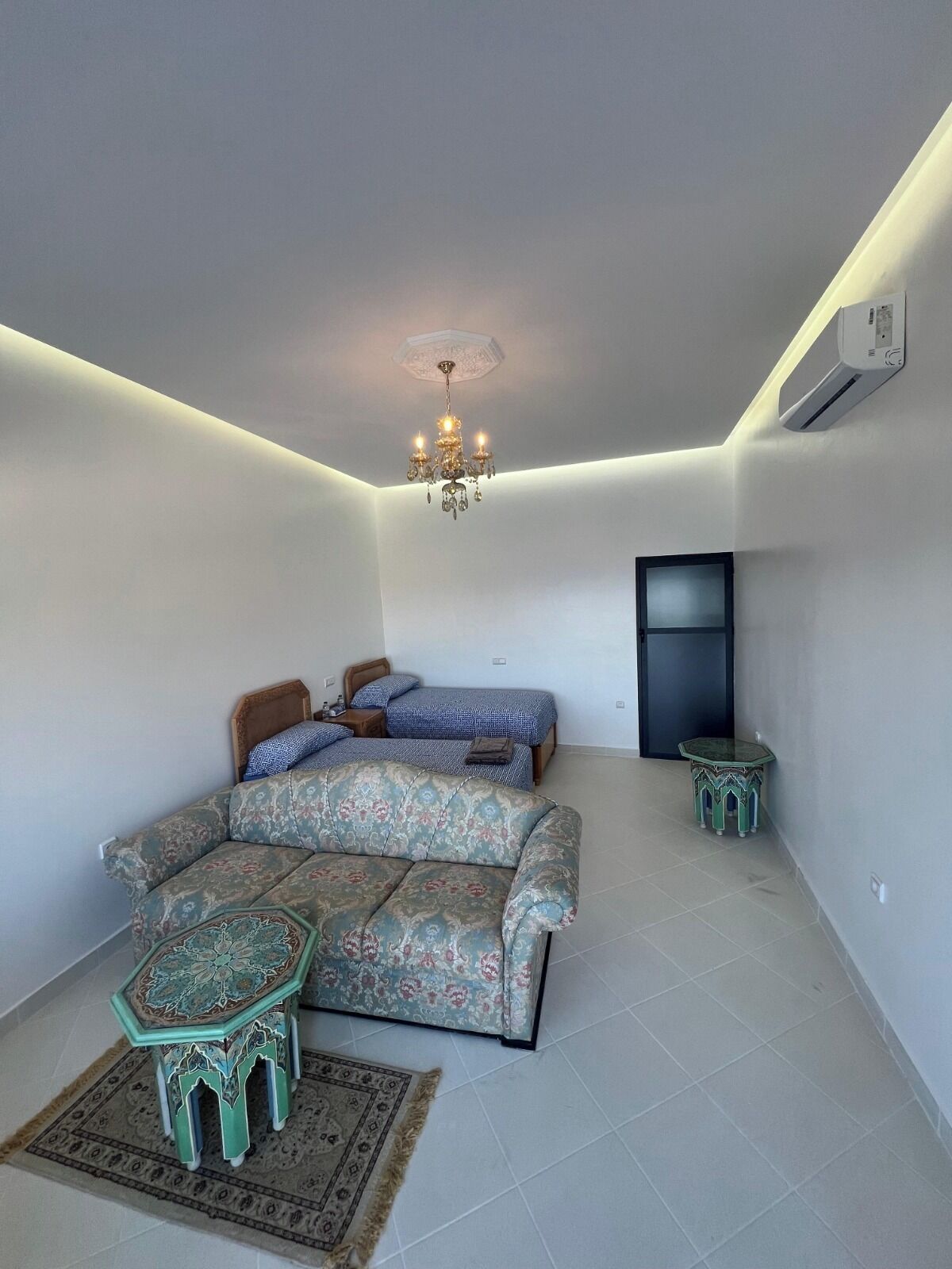 A room with a sofa, two beds, a small table, a chandelier, and an air conditioning unit.