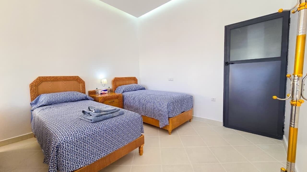 A hotel room with two beds, a nightstand, a lamp, and a door with a security system.