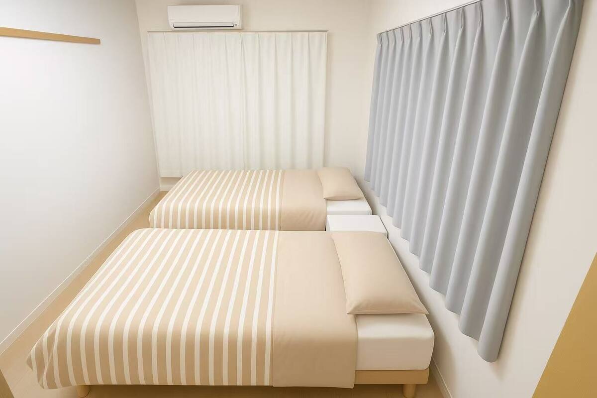 A small, single-room hotel room with two beds, a window with curtains, and an air conditioning unit on the wall.