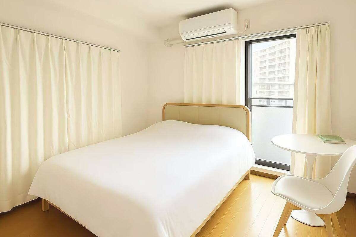 A hotel room with a bed, a round table with a chair, an air conditioning unit, and a window with curtains.