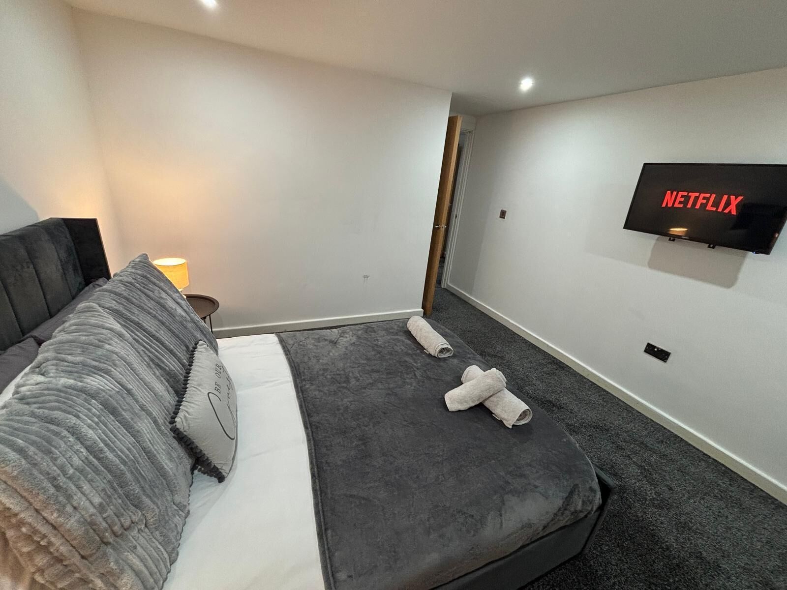 Moderni hotellihuone, jossa on suuri sänky, seinälle asennettu taulutelevisio ja pieni yöpöytä, jossa on lamppu.
