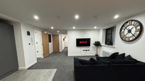 Ett modernt vardagsrum med en svart soffa, en stor väggklocka och en platt-TV som visar ”NETFLIX”.