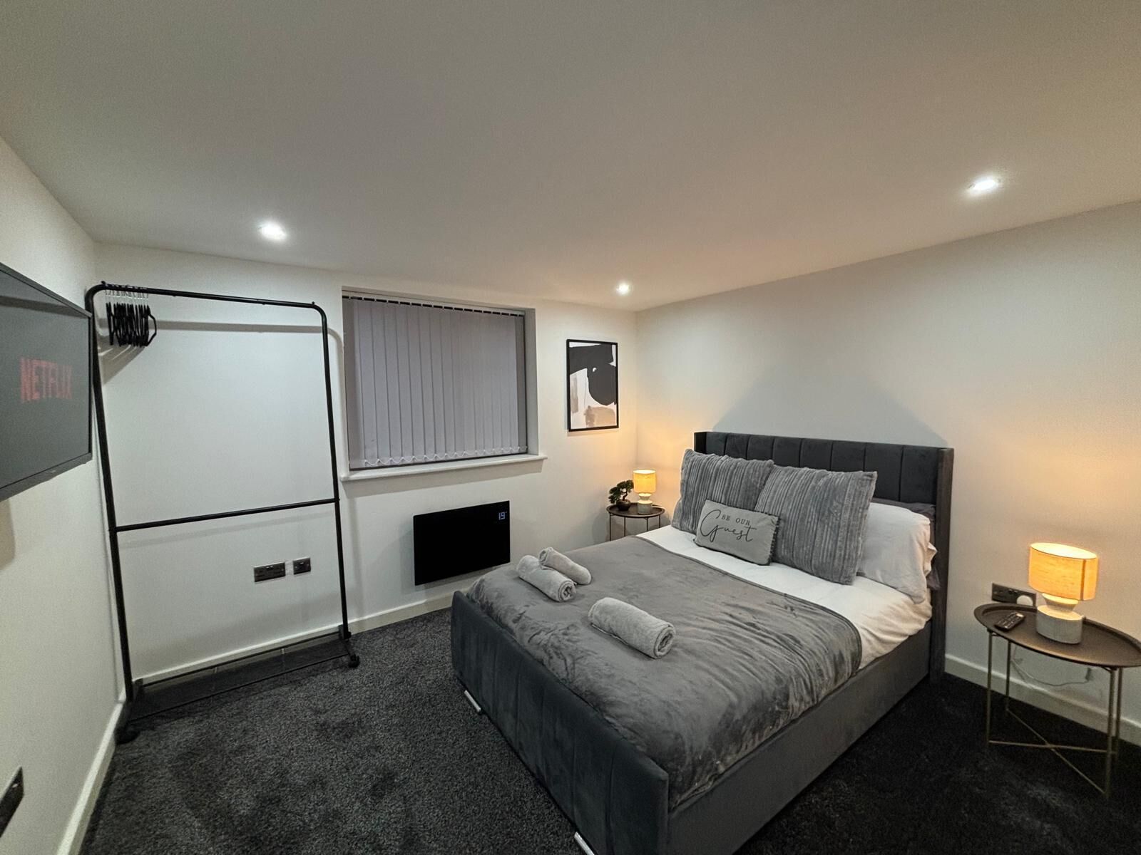 Une chambre d’hôtel moderne dotée d’un grand lit, d’une télévision, d’une fenêtre avec stores, d’une table de chevet avec une lampe et d’un radiateur fixé au mur.