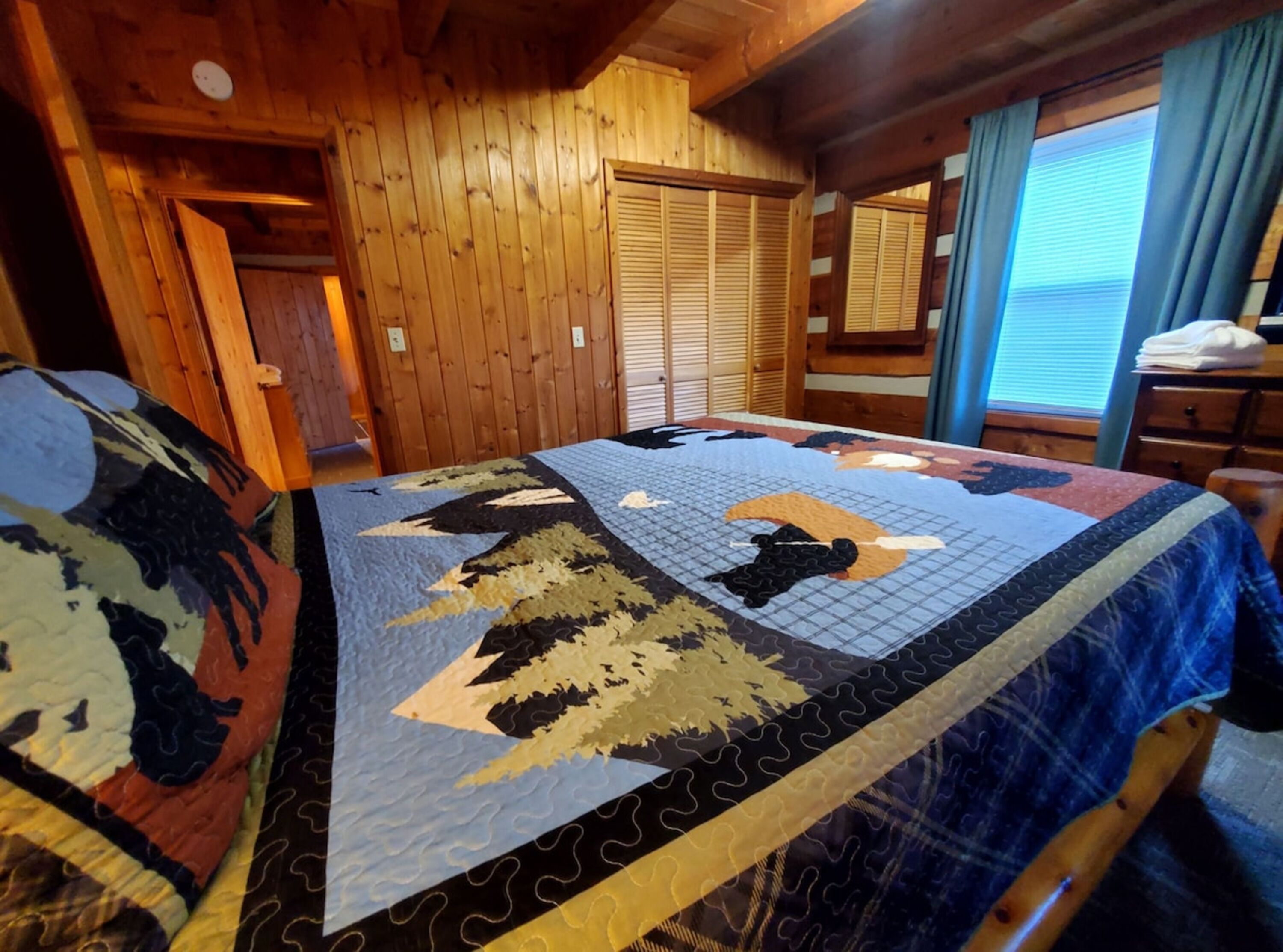 A wooden cabin room with a bed covered in a quilt, wooden walls, a window with curtains, and a wooden door.