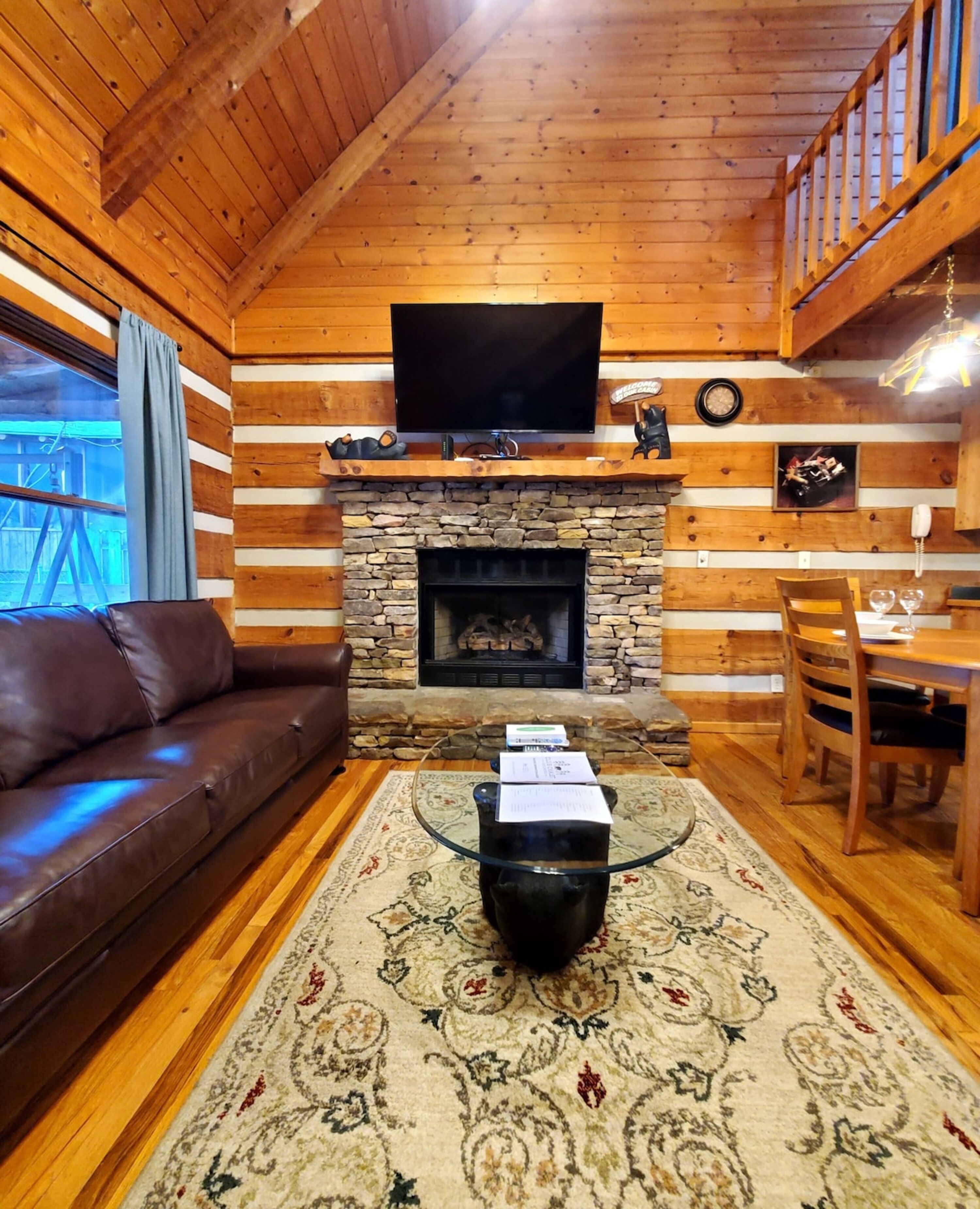 A wooden cabin interior with a stone fireplace, a leather sofa, a glass coffee table, and a dining area with wooden chairs and a table.