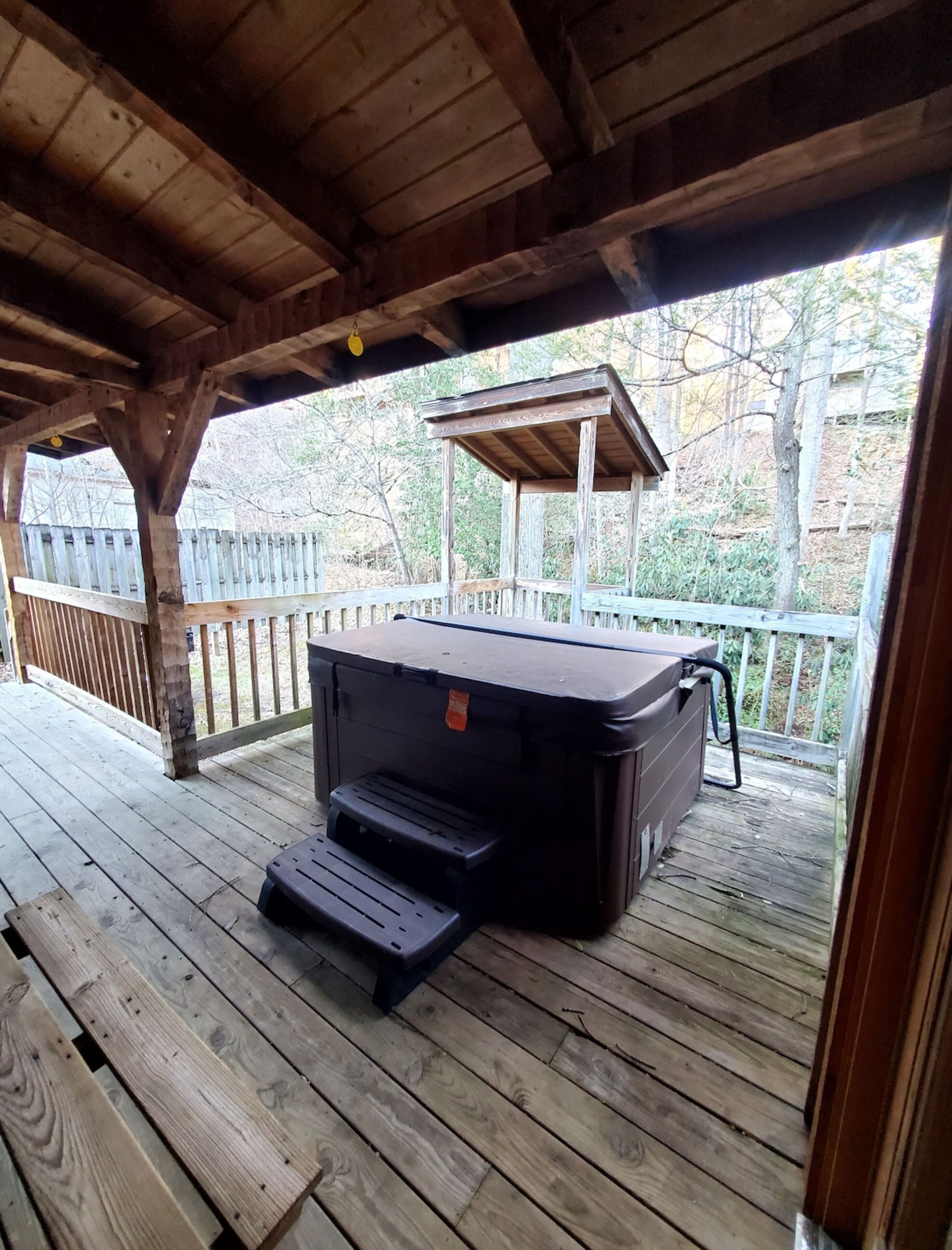 A covered outdoor deck with a hot tub, wooden steps, and a wooden railing.