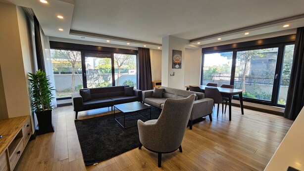 Una sala de estar moderna con un sofá seccional, sillones, una mesa de centro y un comedor con mesa y sillas. Amplias ventanas ofrecen vistas al exterior.