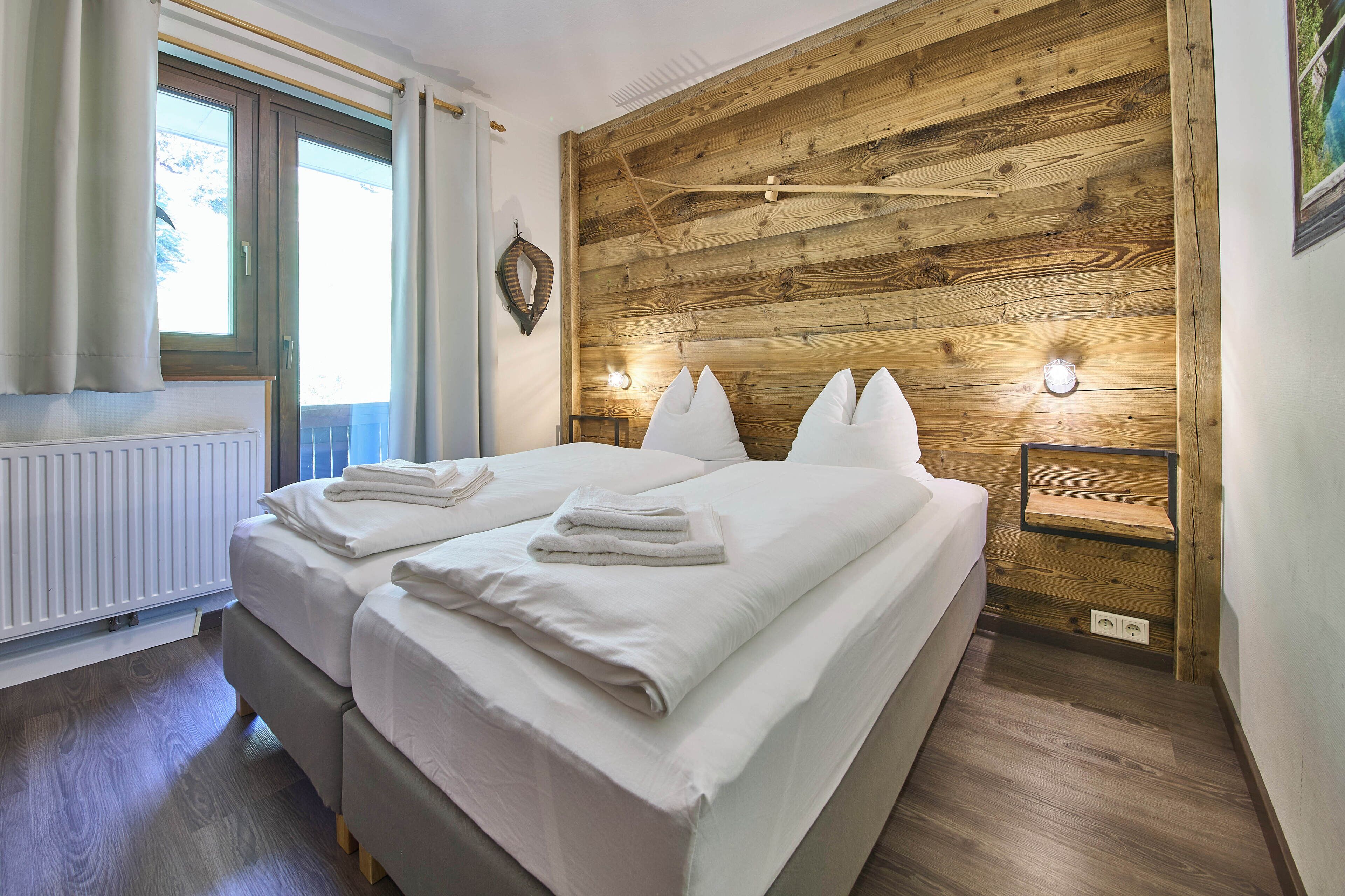 A bedroom with two beds, wooden headboard, bedside lamps, and a window with curtains.