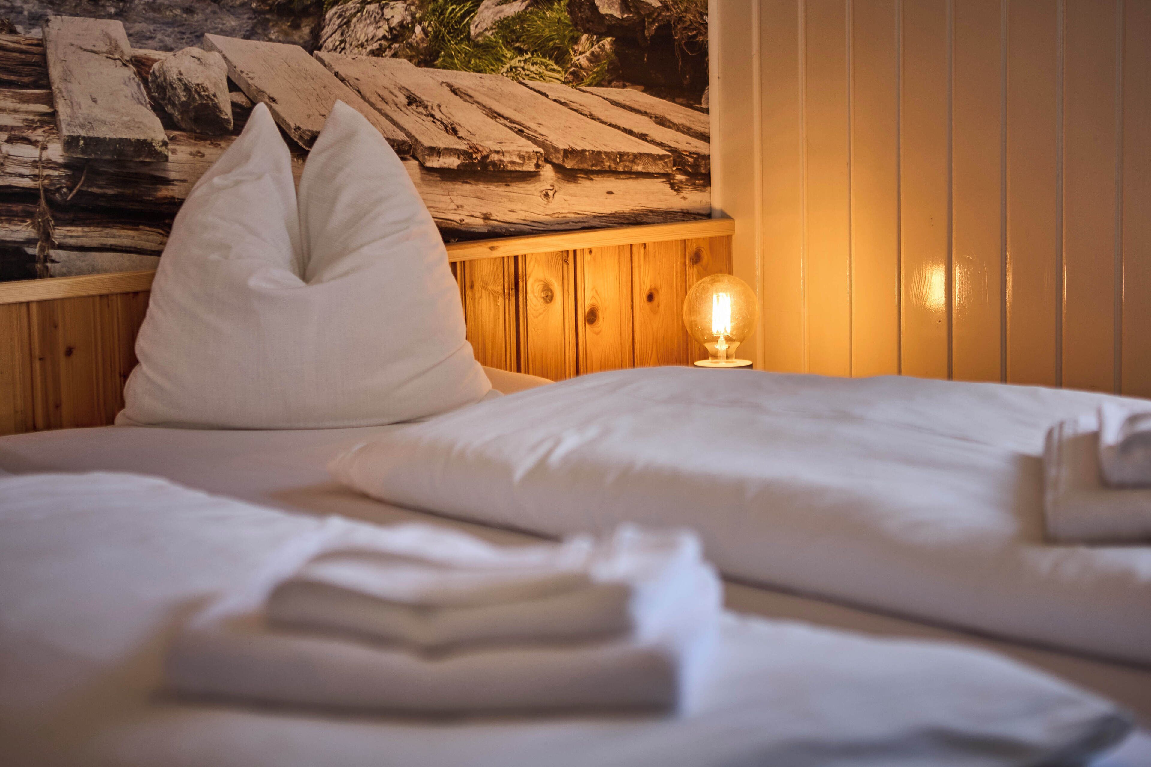 A neatly made bed with white linens and pillows, a wooden headboard, and a wall-mounted lamp.