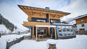 A two-story wooden chalet with a balcony, a glass-enclosed porch, and a snow-covered landscape. - Apartments Enzian (Rauris)