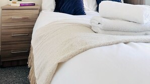 Una cama bien hecha con ropa de cama blanca y azul marino, una manta color beige y una mesita de noche de madera con una planta pequeña y libros.