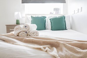 Una cama bien hecha con toallas enrolladas, una manta beige y almohadas verdes.