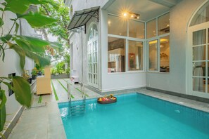 A pool area with a floating basket of fruits, a ladder, and a view of the kitchen through large windows. - Esh Cottages Birch | Hillside 4-BHK Hideaway With Lush Garden, Common Pool, & Gaming Zone (Calangute)