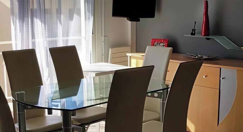 A modern dining area with a glass table and chairs, a television above, and a red vase on a shelf.