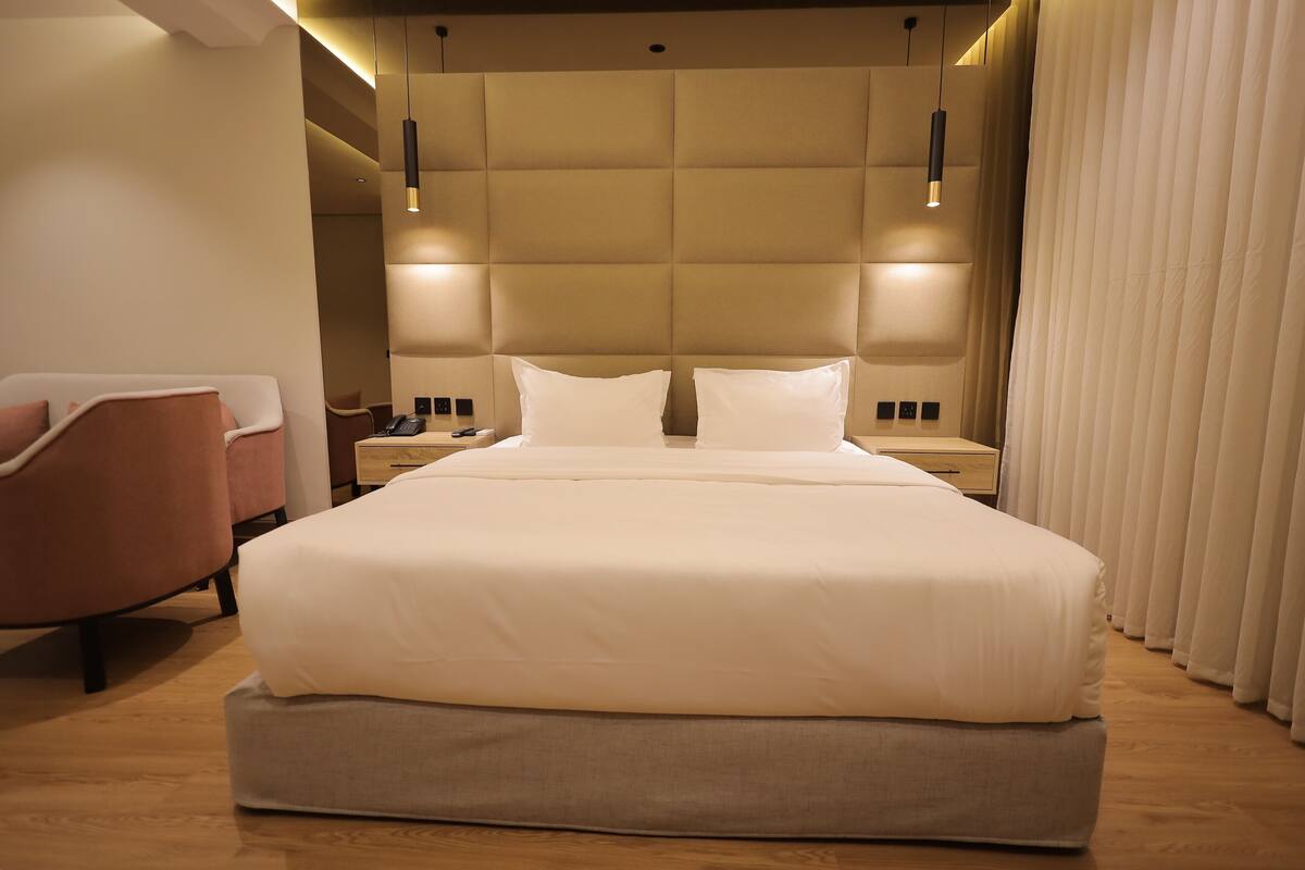 A modern hotel room with a large bed, a bedside table, a chair, and a large window with curtains.