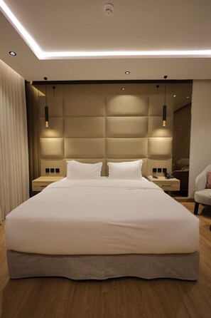 Una habitación de hotel moderna con una cama grande, suelo de madera, cabecera con iluminación integrada, mesitas de noche y un espejo.
