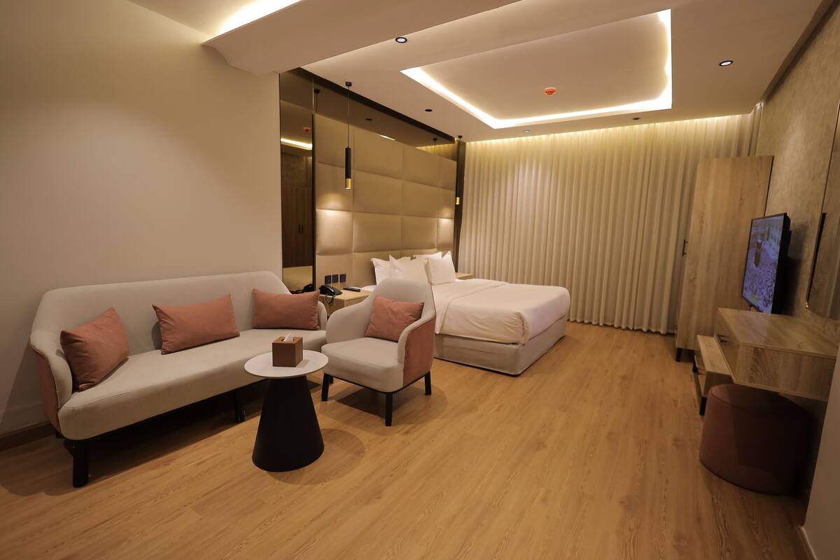 A modern hotel room with a sofa, armchair, and a bed. There is a small table and a TV on a stand.