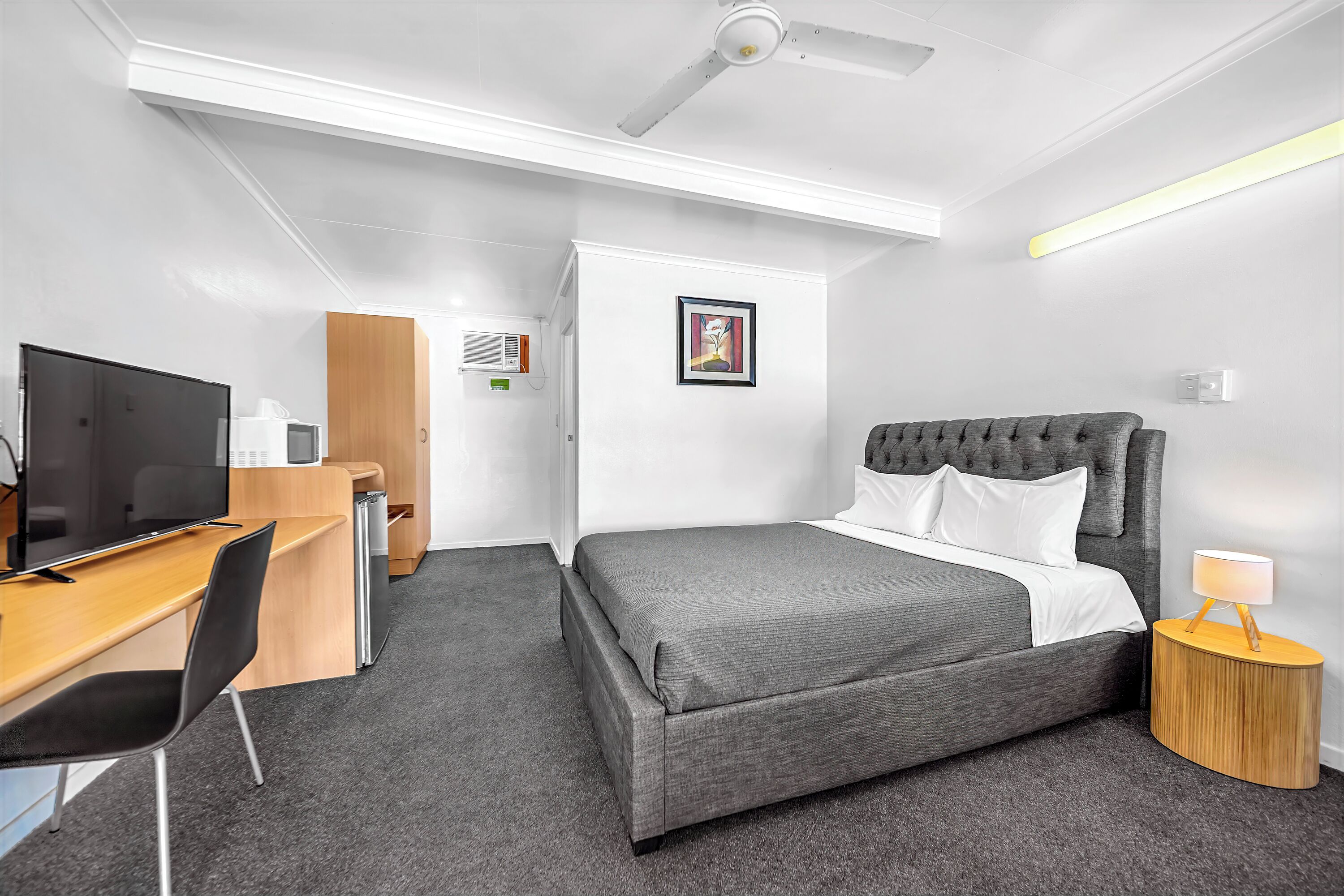A hotel room with a bed, a desk with a television, a chair, a bedside table with a lamp, and a ceiling fan.