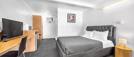 A hotel room with a bed, a desk with a television, a chair, a bedside table with a lamp, and a ceiling fan.