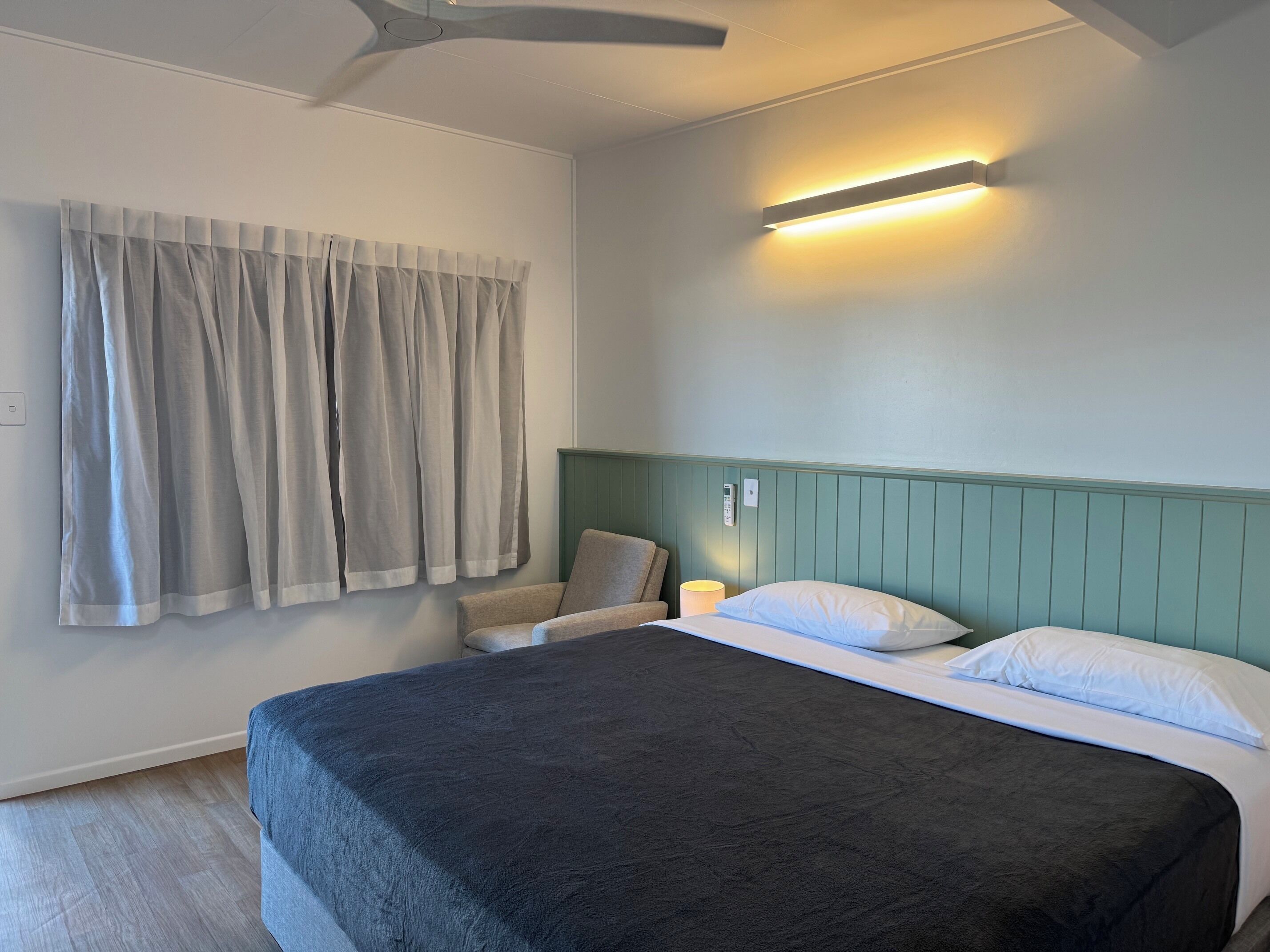 A bedroom with a bed, curtains, a chair, a bedside table with a lamp, and a ceiling fan.