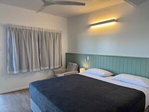 A bedroom with a bed, curtains, a chair, a bedside table with a lamp, and a ceiling fan.