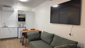 A kitchenette with a microwave, sink, and cabinets, a dining table with chairs, a green sofa, and a flat-screen TV mounted on the wall. - Pearly Shell Motel (Bowen)