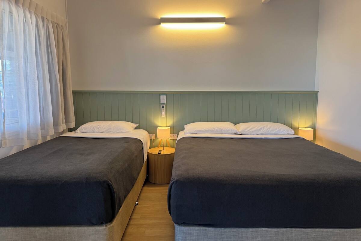 A double room with two beds, a bedside table with a lamp, a wall-mounted light, and a control panel.