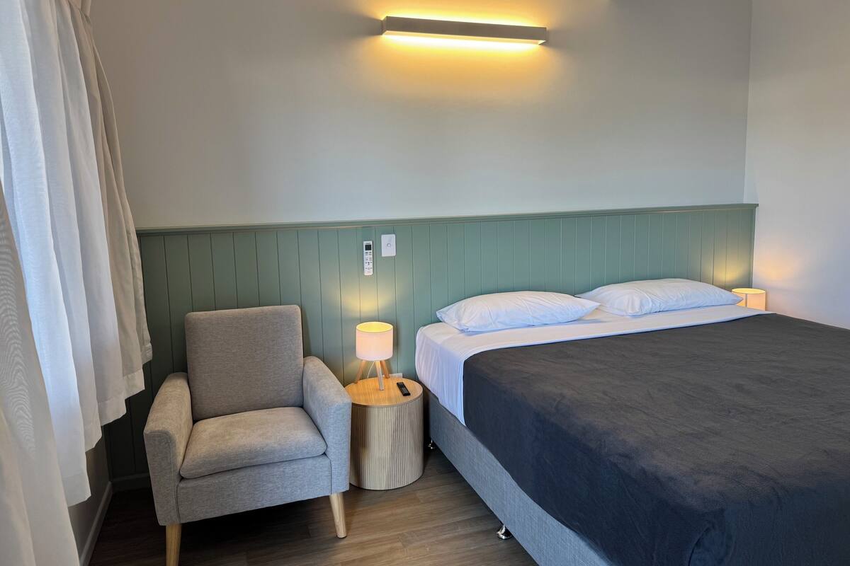 A hotel room with a bed, a chair, a bedside table with a lamp, and a wall-mounted light fixture.