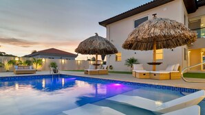 A swimming pool with lounge chairs and thatched umbrellas, surrounded by a house with a balcony. - Ponton 34-g (Oranjestad)