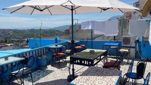 An outdoor terrace with blue and white tiled flooring, multiple tables and chairs, and a large white umbrella providing shade. - hotel sakura (Chefchaouen)