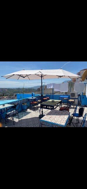 An outdoor terrace with blue and white tiled flooring, multiple tables and chairs, and a large white umbrella providing shade. - hotel sakura (Chefchaouen)