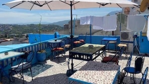 Une terrasse extérieure avec un sol carrelé bleu et blanc, de nombreuses tables et chaises, et un grand parasol blanc offrant de l’ombre.