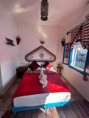 Une chambre à coucher avec un lit, une table de chevet, une lampe fixée au mur et une fenêtre avec des rideaux.