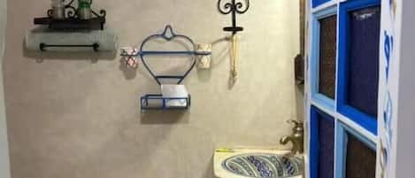 A small bathroom with a toilet, a sink, and a shower. There are shelves with toiletries and a towel rack.