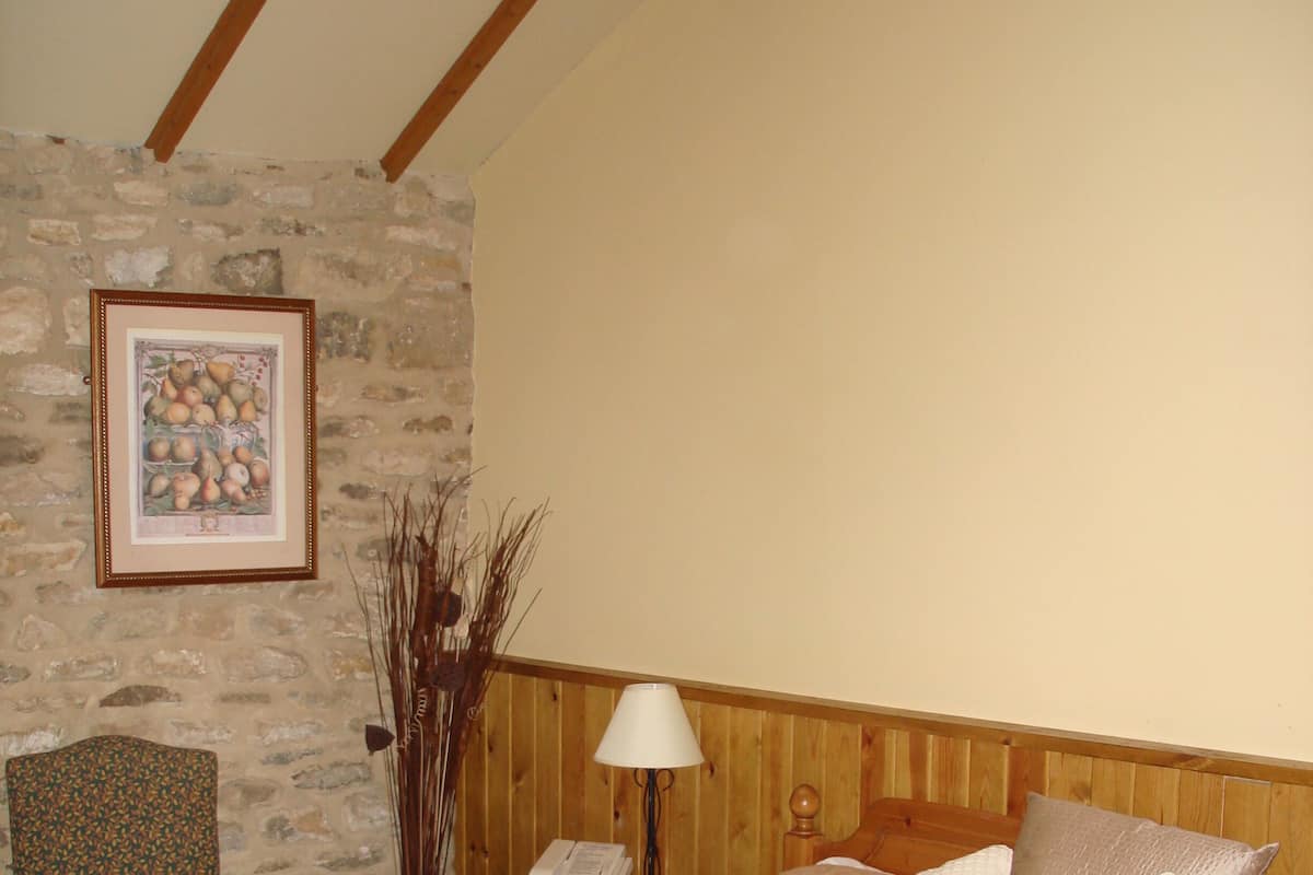 A bedroom with a stone wall, a wooden headboard, a bedside table with a lamp, a telephone, and a floral pillow.