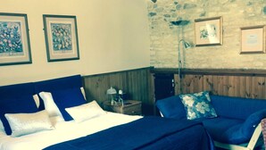 Une chambre avec un grand lit, deux petits lits, des poutres en bois, un mur en pierre, des tableaux encadrés et une table de chevet.