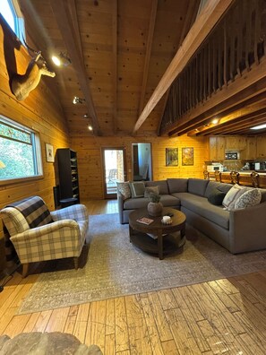A cozy cabin interior with a wooden ceiling, a fireplace, a sofa, a chair, and a dining area.