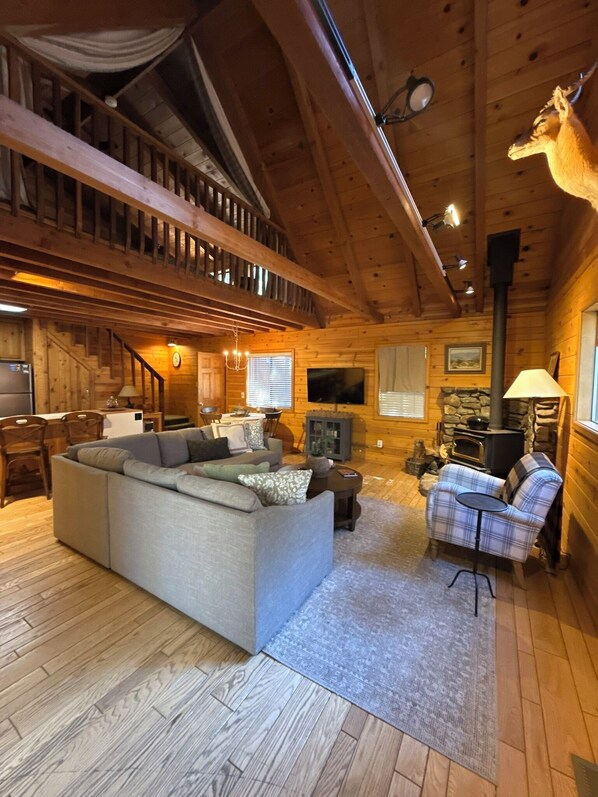 A log cabin interior with a large sofa, a fireplace, a dining area, and a staircase leading to an upper level.
