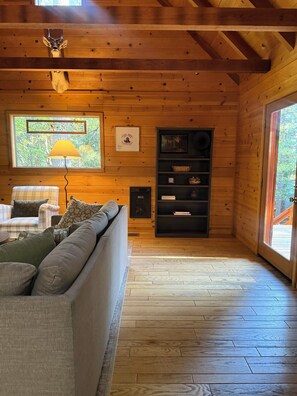 A wooden cabin interior with a sofa, a lamp, a fireplace, and a window with a view of trees.